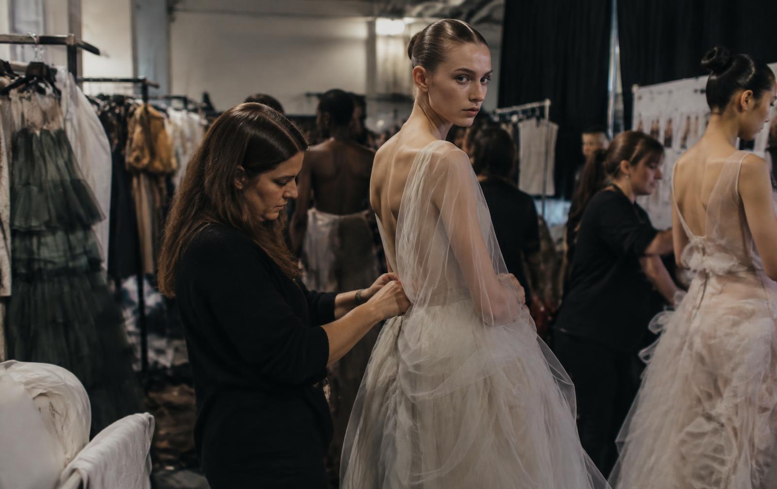 Dressers preparing models in pale layered dresses backstage beside clothing racks before a fashion show