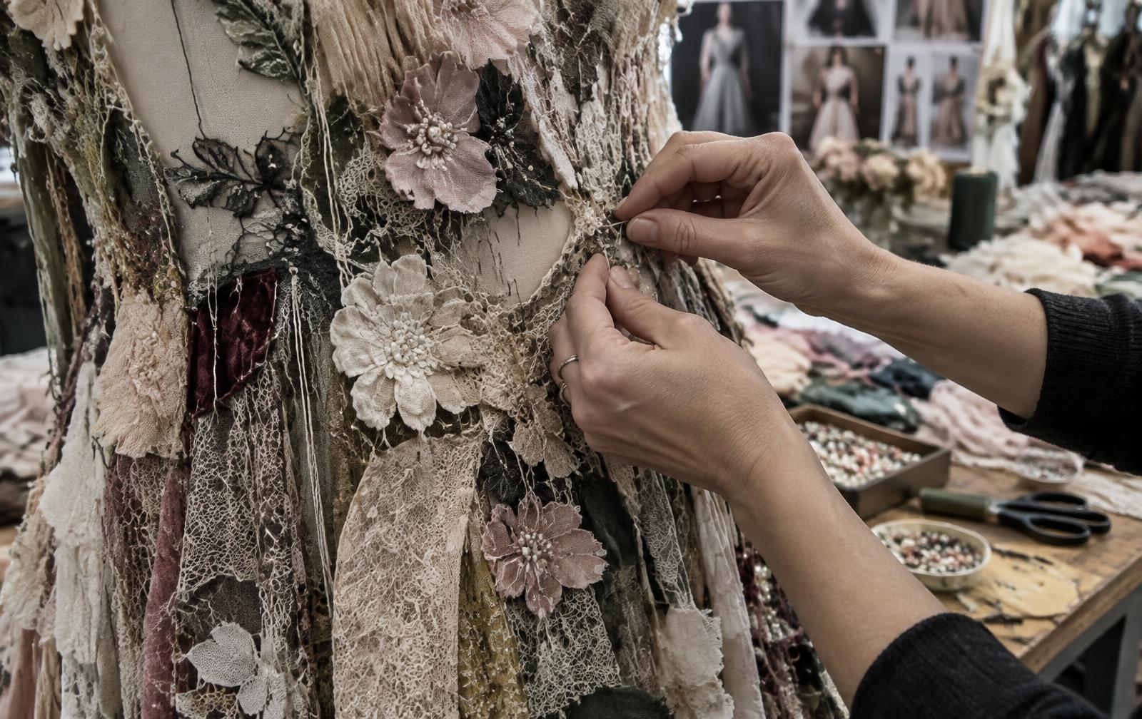 Close view of hands stitching raised fabric flowers and embroidered stems onto textured cloth in a workroom