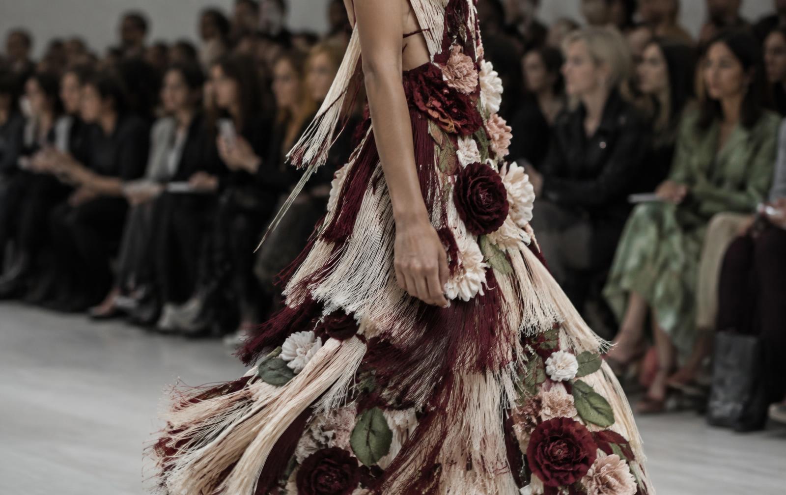 Close runway-side view of a moving dress with burgundy dimensional flowers, soft fringe and textured panels