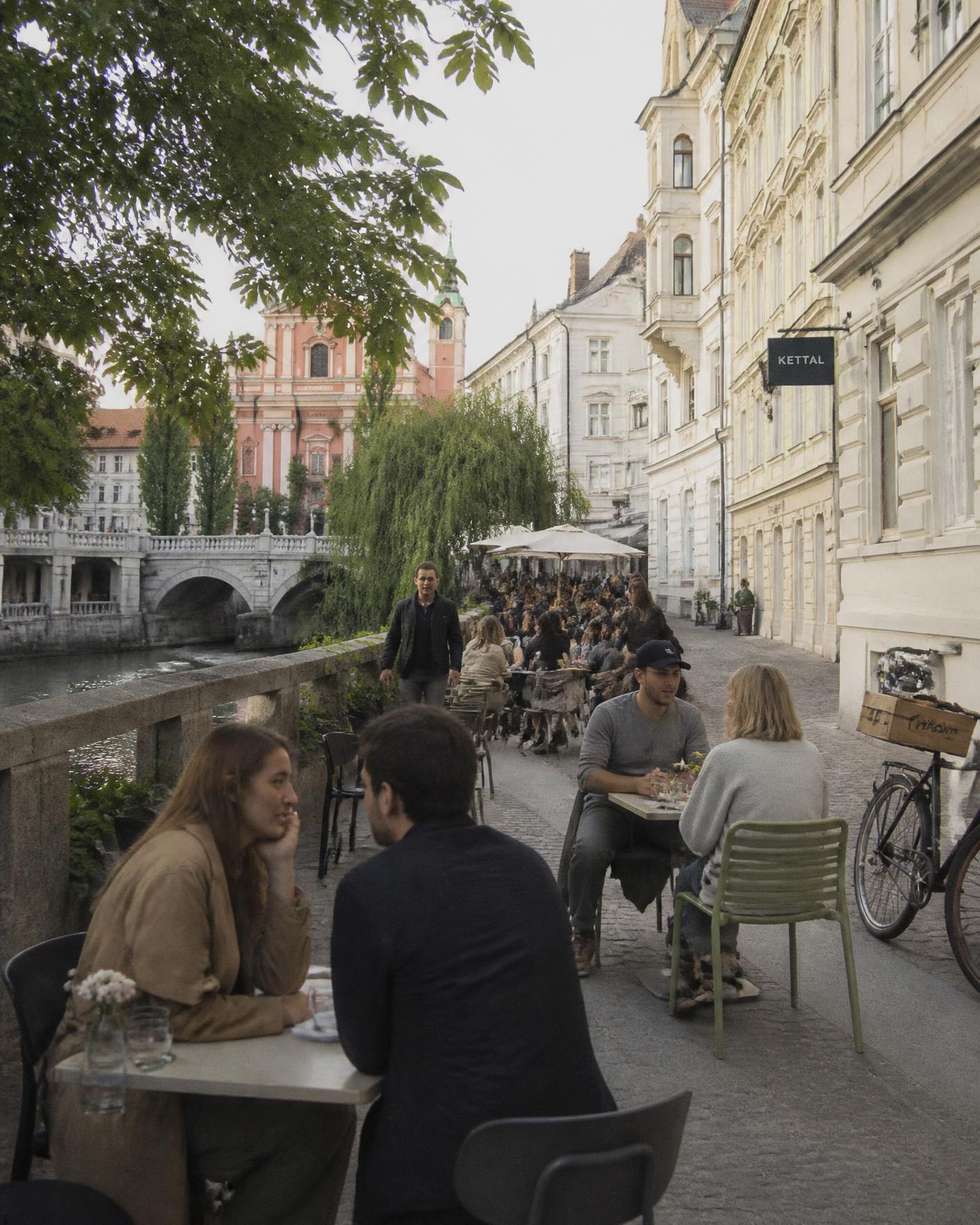 A riverside café terrace in Ljubljana with pastel baroque facades and a stone bridge behind