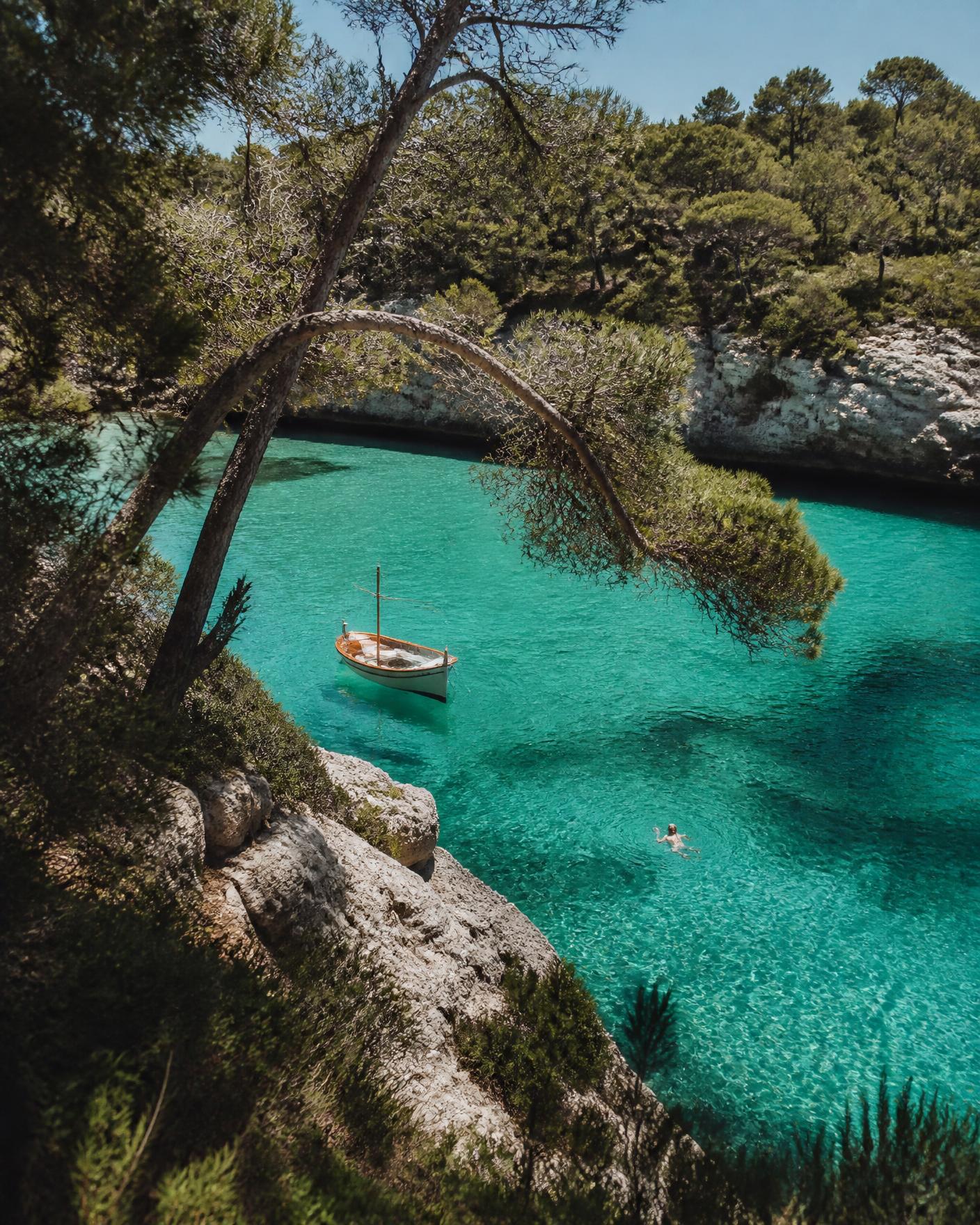 A turquoise limestone cove in Menorca with pine trees and a small wooden boat