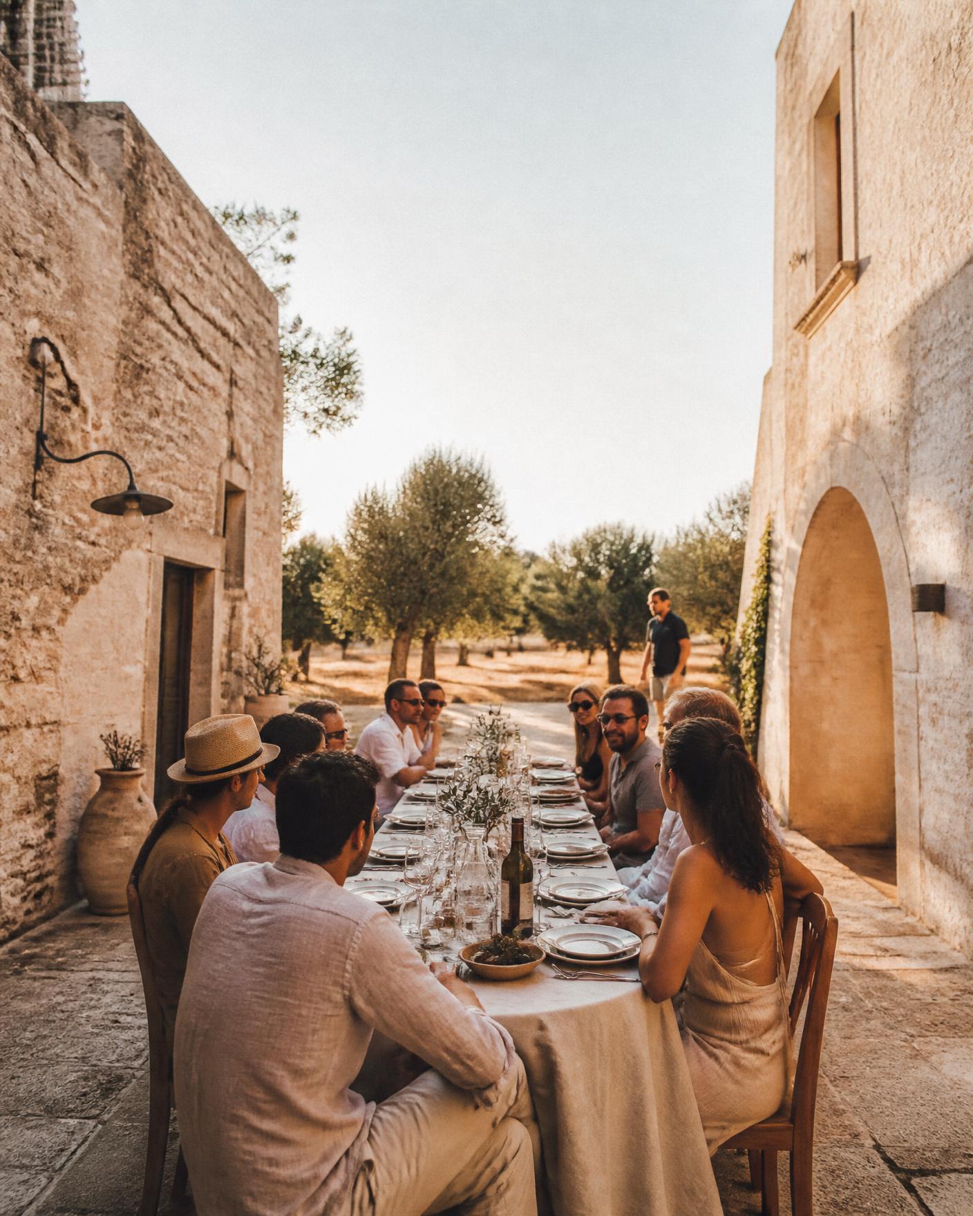 A long lunch table set in the courtyard of a Puglian masseria at golden hour with olive trees beyond