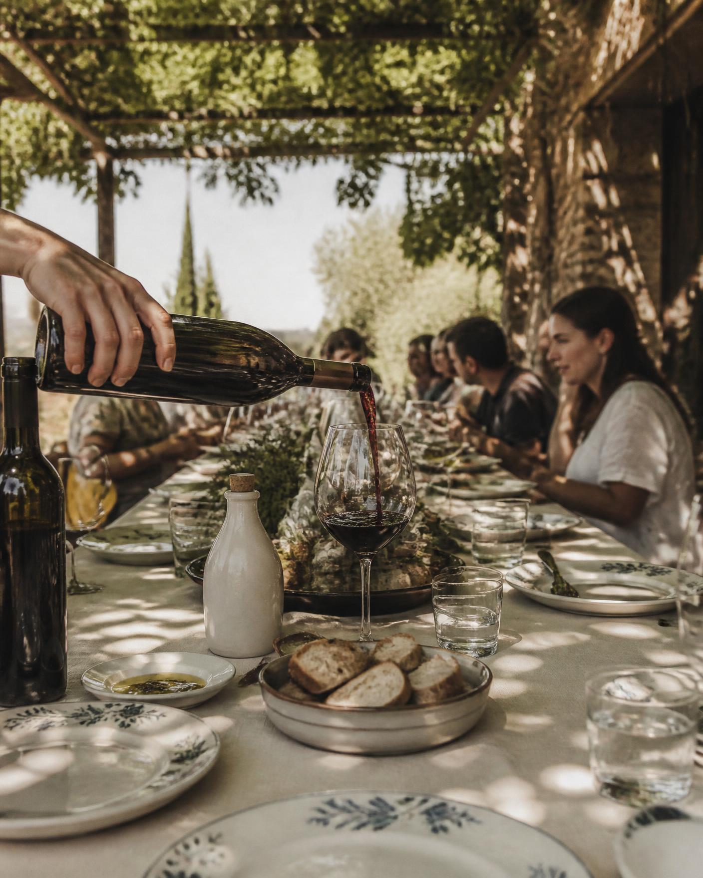 Hands pouring red wine at a long lunch table under a pergola in the Tuscan countryside